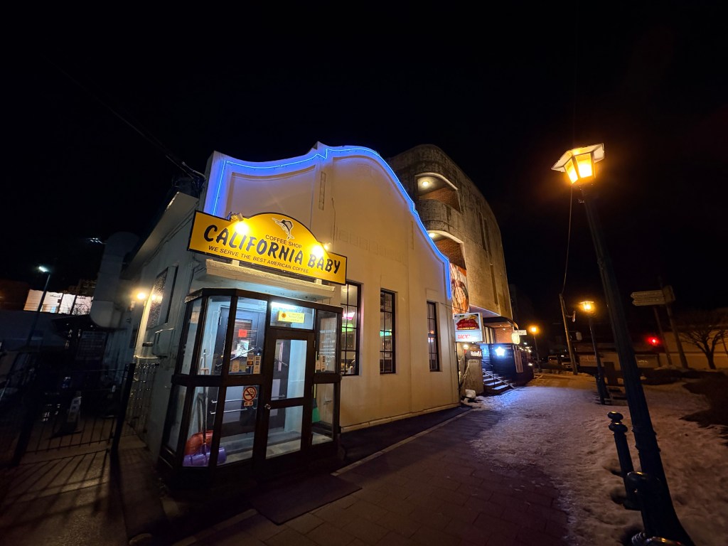 Exterior of California Baby restaurant in Hakodate at night, featuring a bright yellow sign and blue neon lights on a vintage white building.