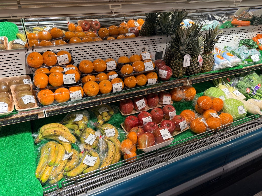 Brightly lit grocery shelves filled with fresh Hokkaido vegetables like green onions and radishes, and fruits like oranges and pineapples.