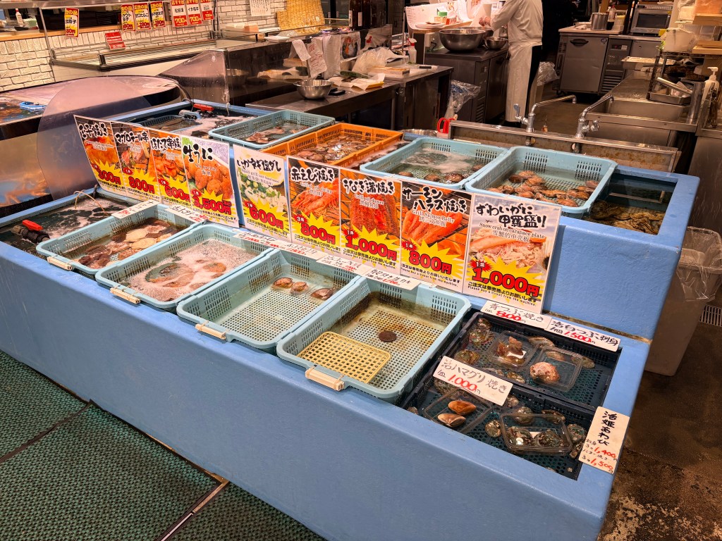 Blue water tanks filled with live seafood and colorful menu signs in a market setting.