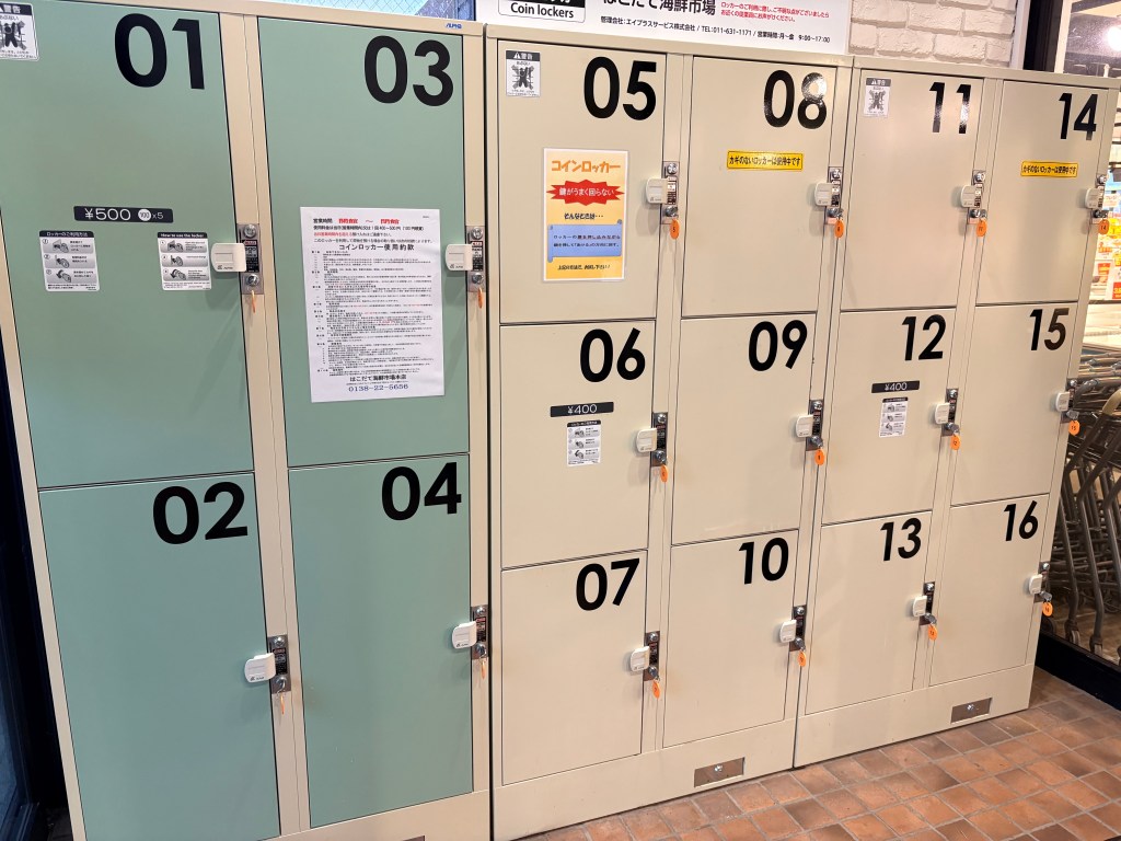 A row of silver coin-operated lockers in various sizes, including large ones suitable for suitcases, located near the market entrance.