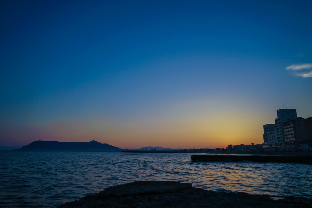 A scenic coastal view of the Tsugaru Strait at dusk in Hakodate, with the dark silhouette of Mt. Hakodate on the horizon and hotels along the shoreline under a deep blue and orange sunset sky.