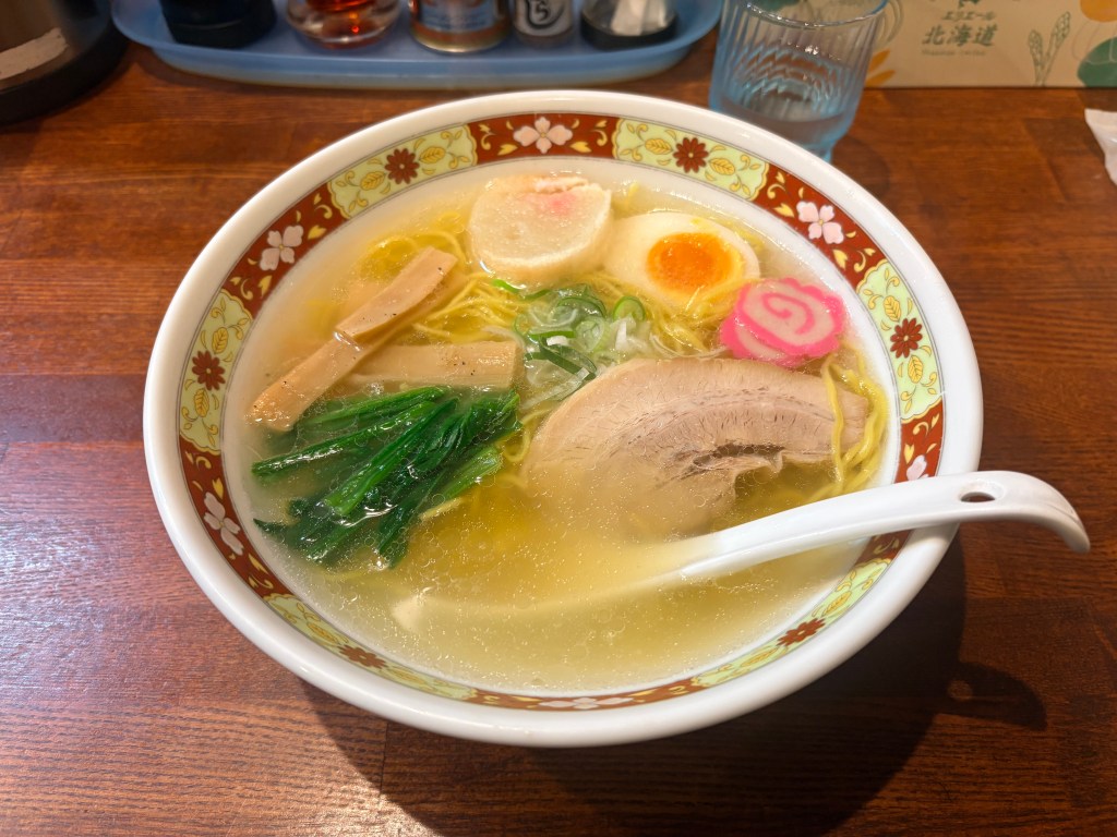 A bowl of Hakodate Shio Ramen with clear broth, pork slices, a soft-boiled egg, and naruto fish cake.