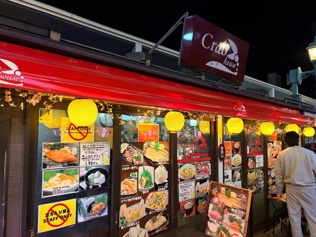 The exterior of "Club House" at Daimon Yokocho, decorated with glowing yellow lanterns and colorful food photos on the window.