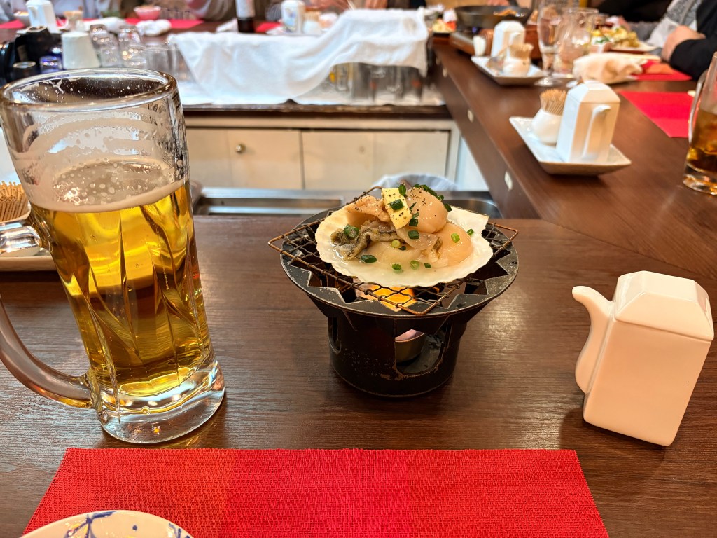 A large scallop being grilled in its shell with butter and green onions over a small portable flame on a bar counter.