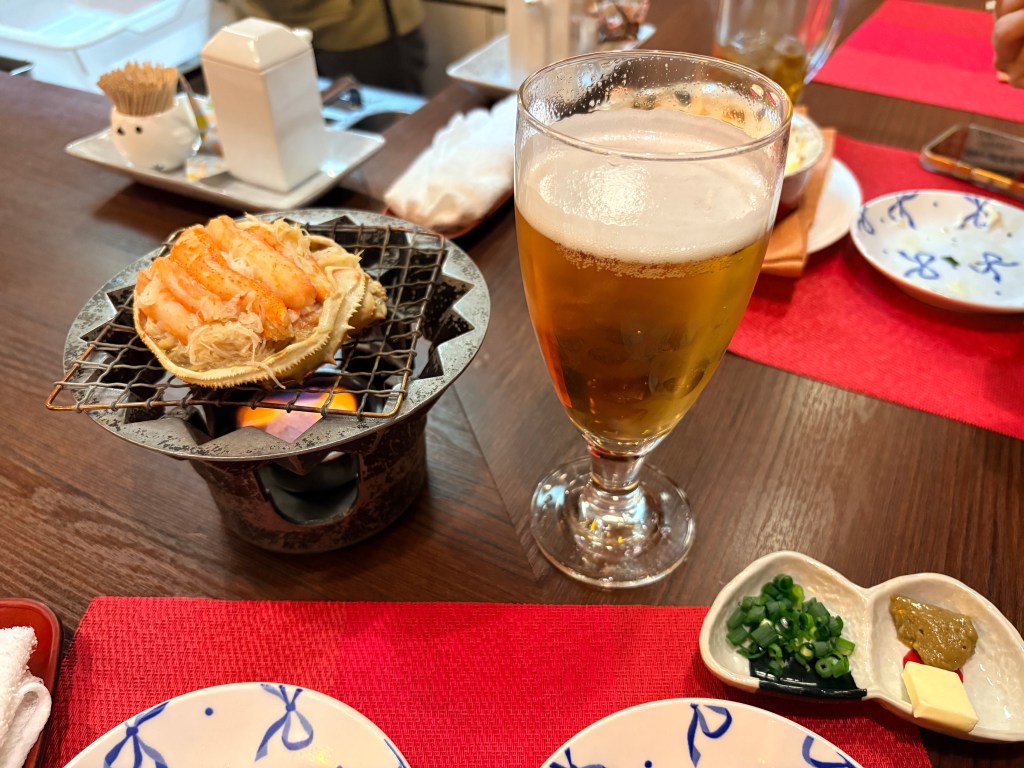 A crab shell filled with crab meat and miso being heated over a flame, served with a glass of beer and condiments.