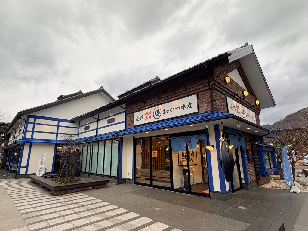 The exterior of Marukatsu Suisan in Hakodate's Bay Area, featuring a traditional Japanese-style building with a large iconic tuna model hanging near the entrance.