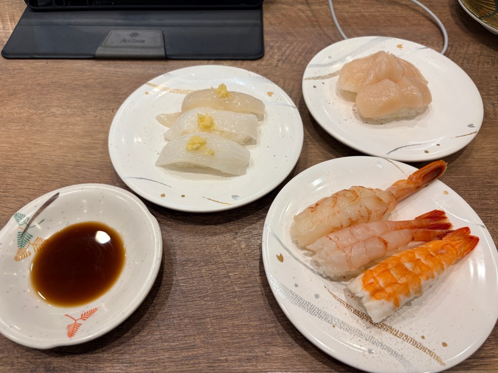 A variety of fresh sushi on a table, including squid topped with ginger, a large scallop, and three types of shrimp, all served on clean white plates.