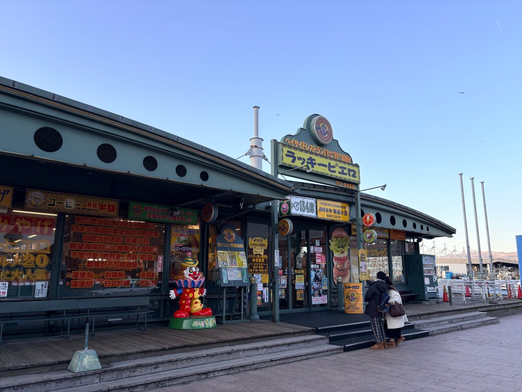 Exterior view of the green Lucky Pierrot restaurant with a colorful clown statue at the entrance and people standing on a wooden deck.