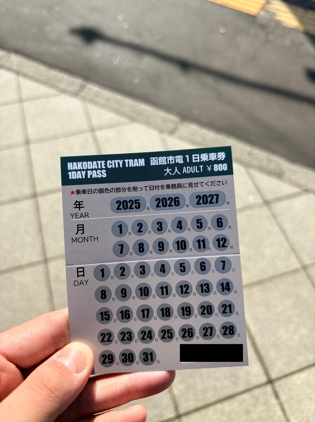 A person's hand holding a physical Hakodate City Tram 1-Day Pass. The card is grey and green, featuring rows of silver scratch-off circles for the year, month, and day. The top of the card displays the price of 800 yen for adults.