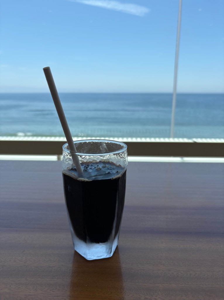 An iced coffee on a cafe table, with a breathtaking view of the deep blue Tsugaru Strait stretching to the horizon through a massive panoramic window.