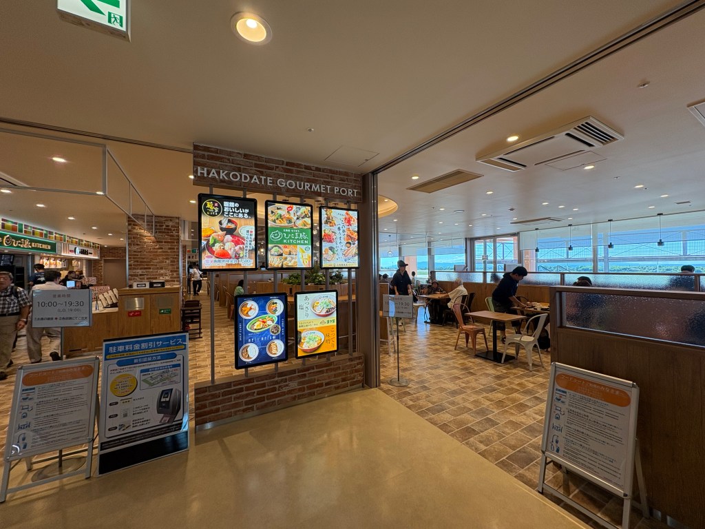 A wide, panoramic view of the bright and clean food court area on the 3rd floor of Hakodate Airport, showing numerous wooden tables and various restaurant stalls in the background.