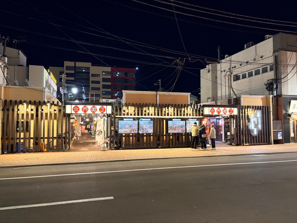 A wide night shot of the illuminated entrance to Daimon Yokocho Food Stall Village in Hakodate, featuring bright signs and wooden fences along the street.