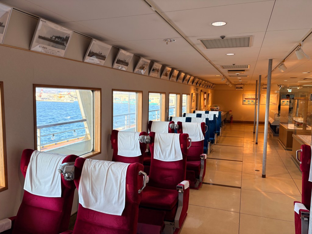 Rows of retro red fabric-covered double seats inside the Mashu Maru cabin, with blue sea visible through the windows.