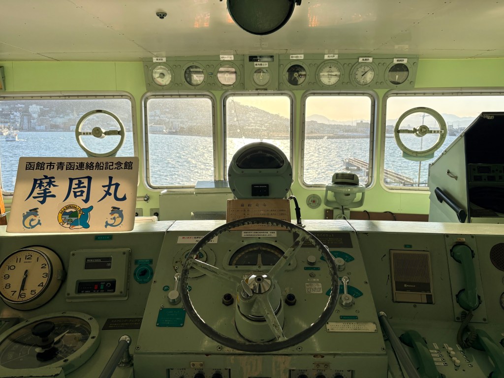 Inside the Wheelhouse (Bridge) of the Mashu Maru. A large wooden steering wheel is in the center, looking out through the windows at the clear blue waters of Hakodate Harbor and Mount Hakodate in the distance.