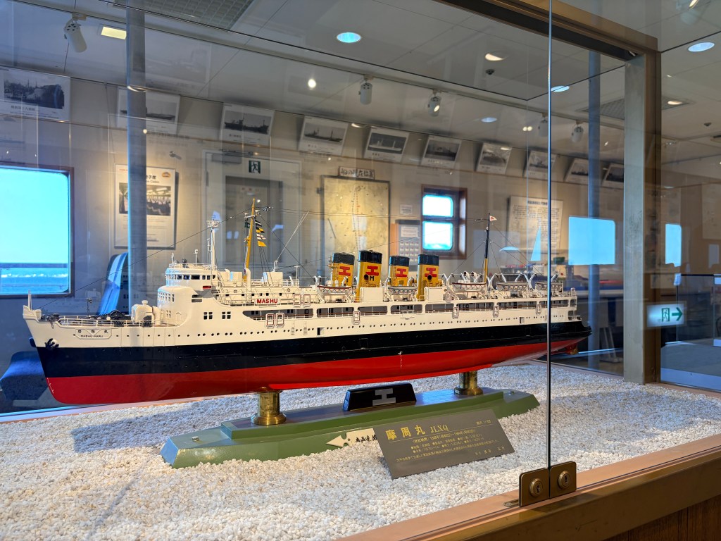 A detailed scale model of the first Mashu Maru in a glass case, showing its iconic yellow funnels.