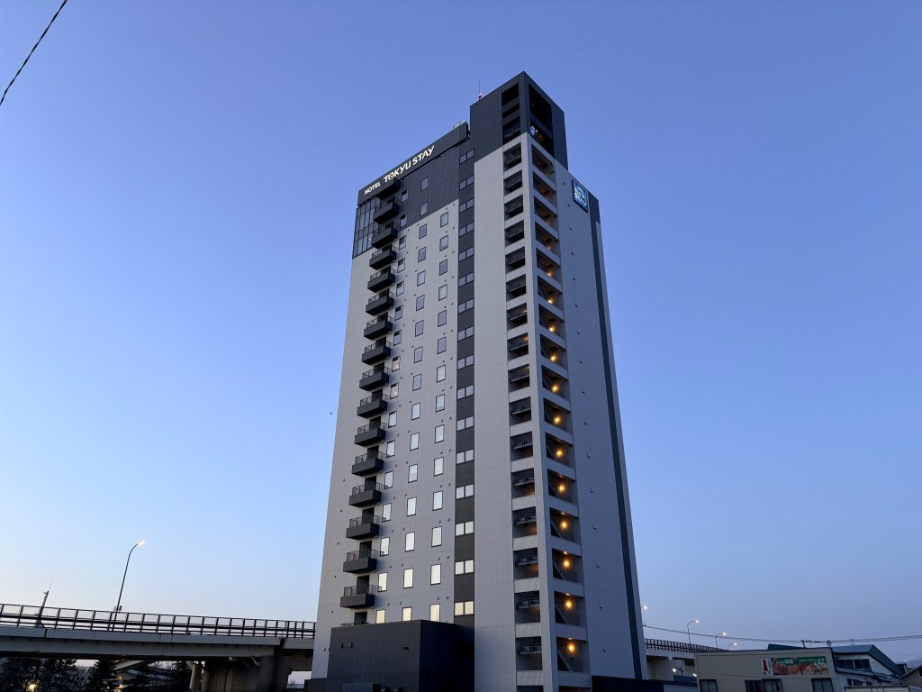 A daytime photograph of the modern, multi-story facade of the Tokyu Stay Hakodate Asaichi Akari no Yu building under a clear blue sky.