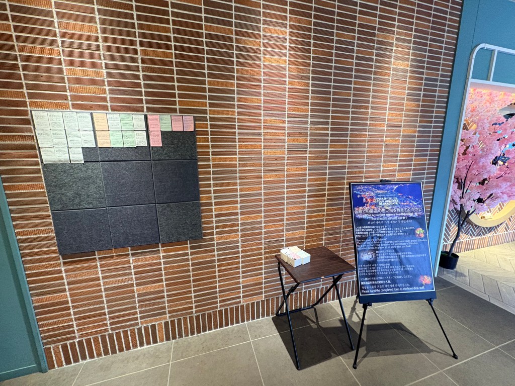 A brick-textured wall in the hotel lobby featuring a board with colorful handwritten guest feedback cards and a nearby informational sign on an easel.