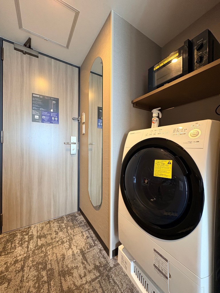 A close-up shot within a hotel room showing a white washer/dryer unit positioned below a built-in shelf holding a silver microwave oven.