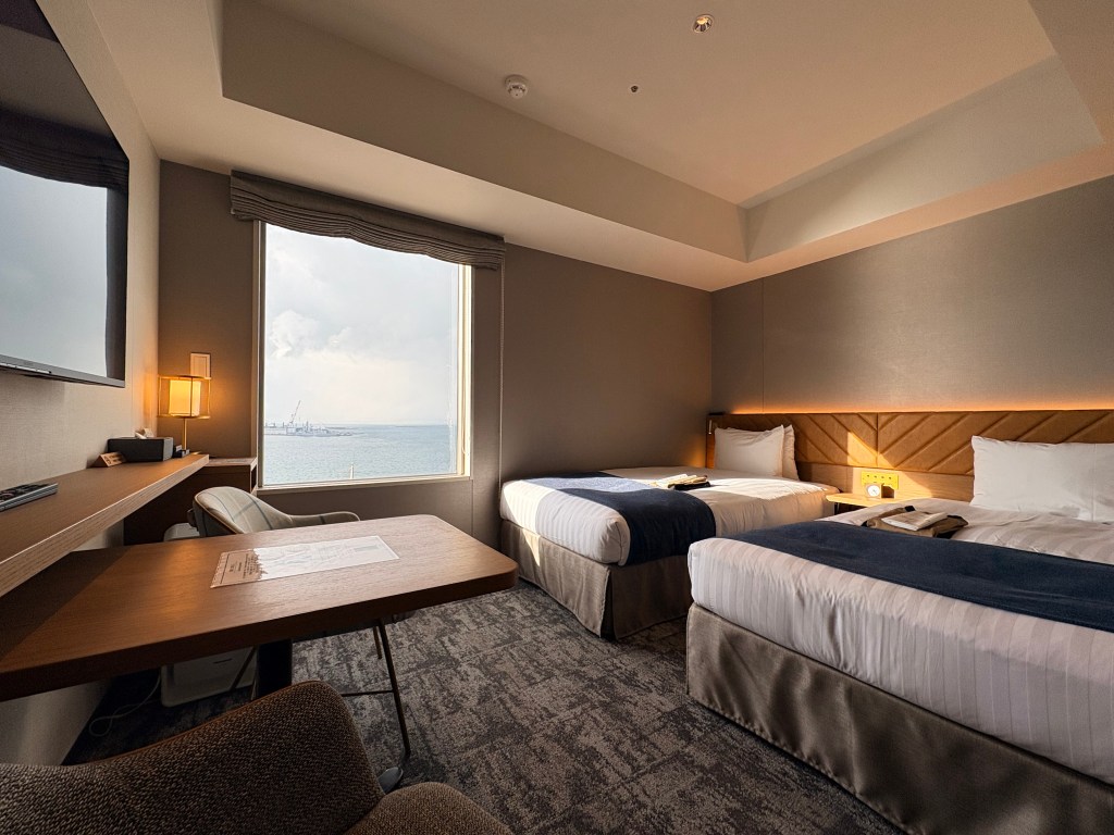 A wide-angle view of a modern hotel twin room featuring two beds with blue runners, a wooden work desk, and a large window overlooking the ocean.
