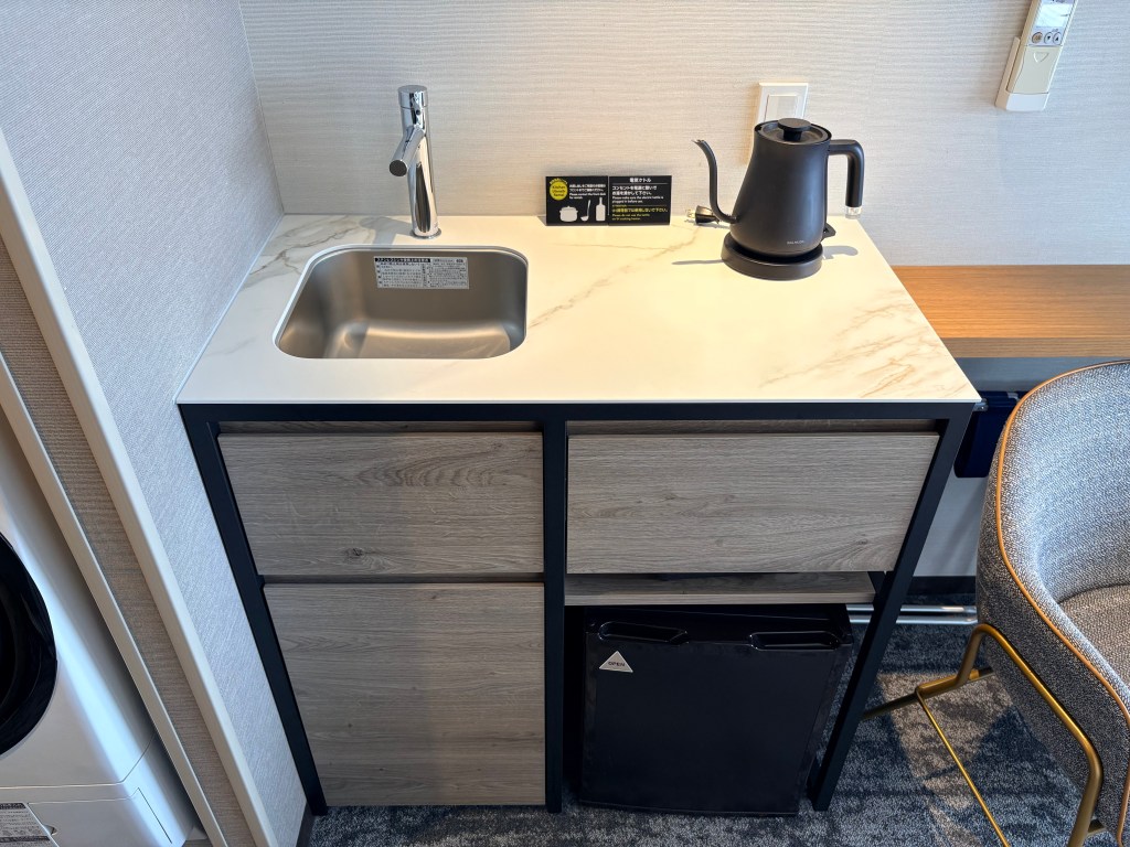 A photograph of a small, modern hotel kitchenette featuring a stainless steel sink, a single IH burner, an under-counter refrigerator, and upper cabinets.