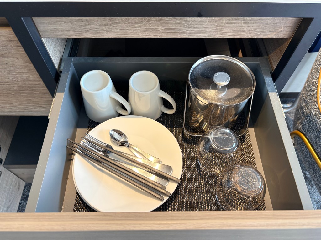 An open drawer in the kitchenette containing sets of ceramic plates, bowls, mugs, and cutlery neatly arranged for guests.