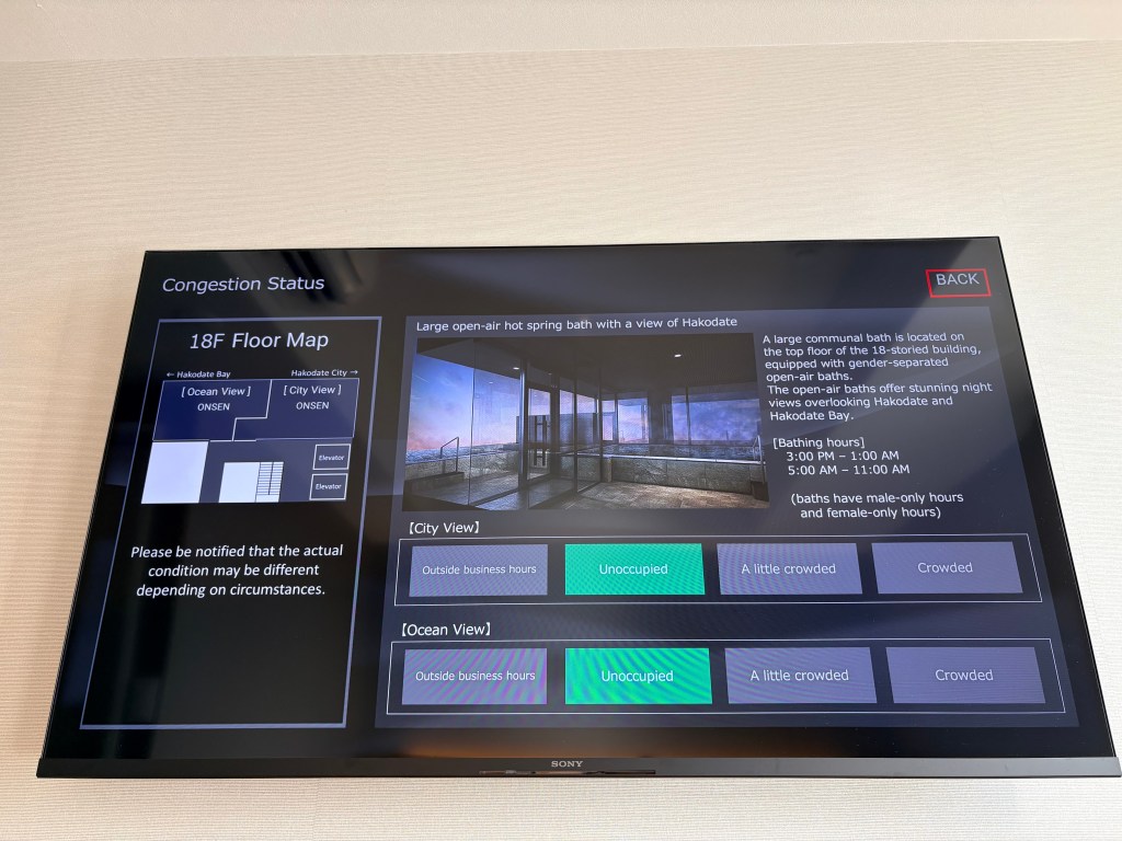A flat-screen TV displaying the hotel's information menu, specifically showing the real-time occupancy levels of the 18th-floor public baths.
