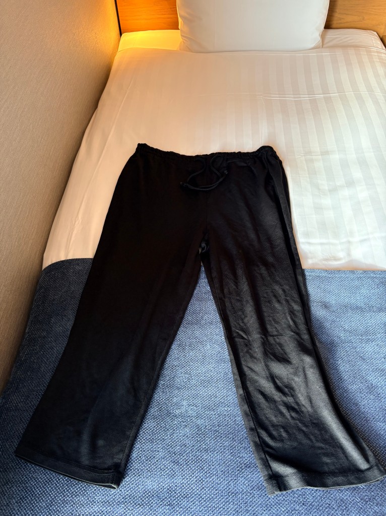 A pair of black, loose-fitting drawstring pajama pants laid out on a white hotel bed.