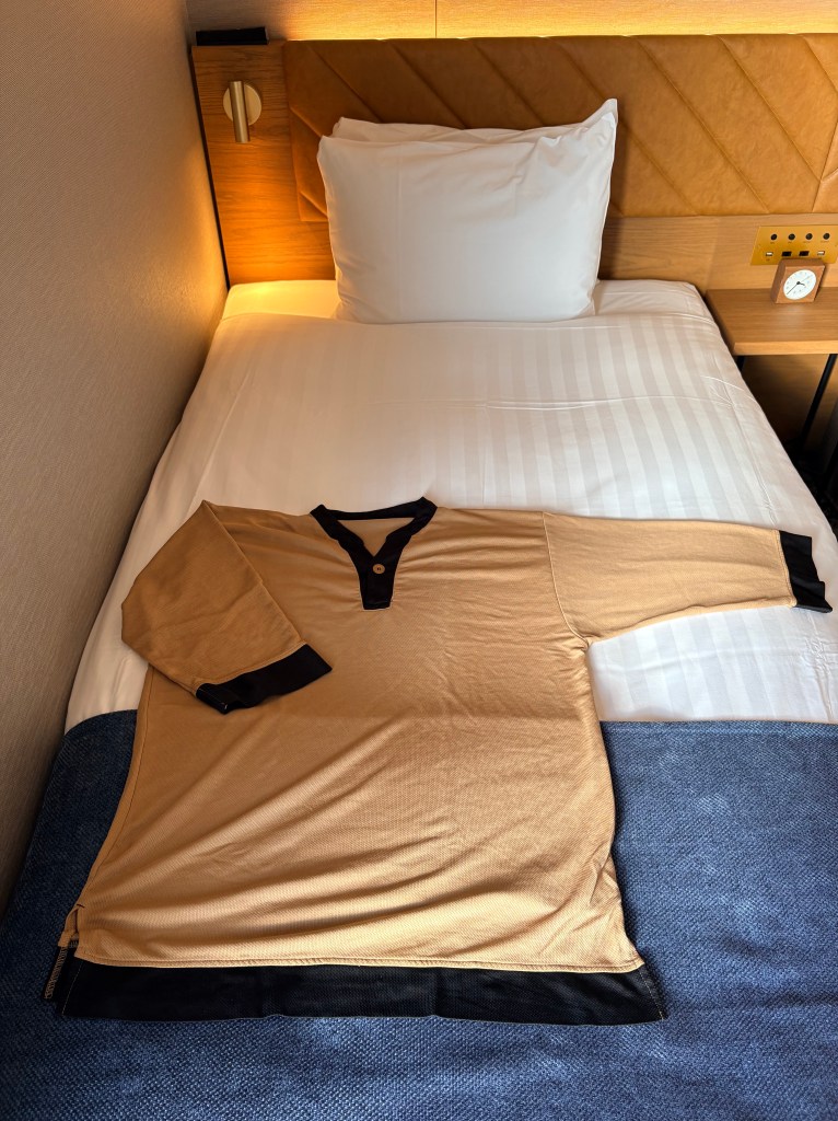 A tan-colored V-neck room wear shirt with dark trim laid out on a white hotel bed.