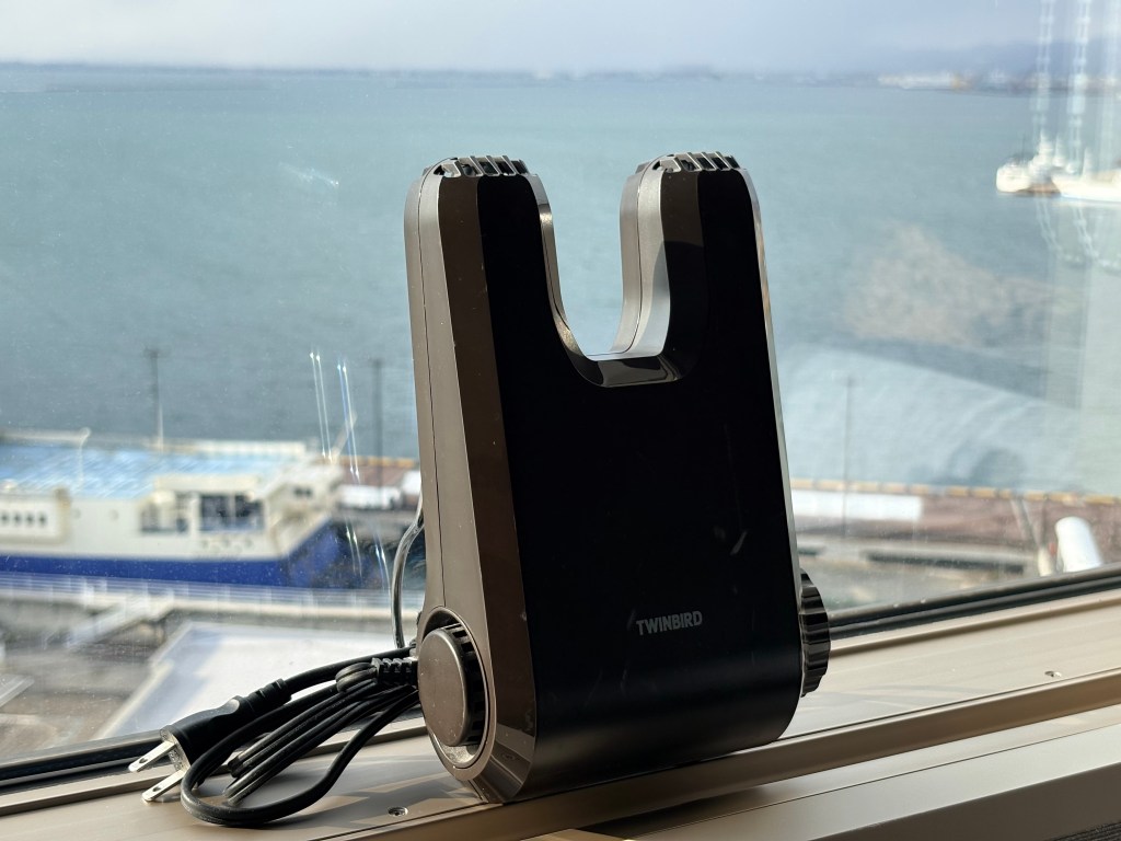 A black Twinbird electric shoe dryer placed on the windowsill with a view of the Hakodate harbor in the background.