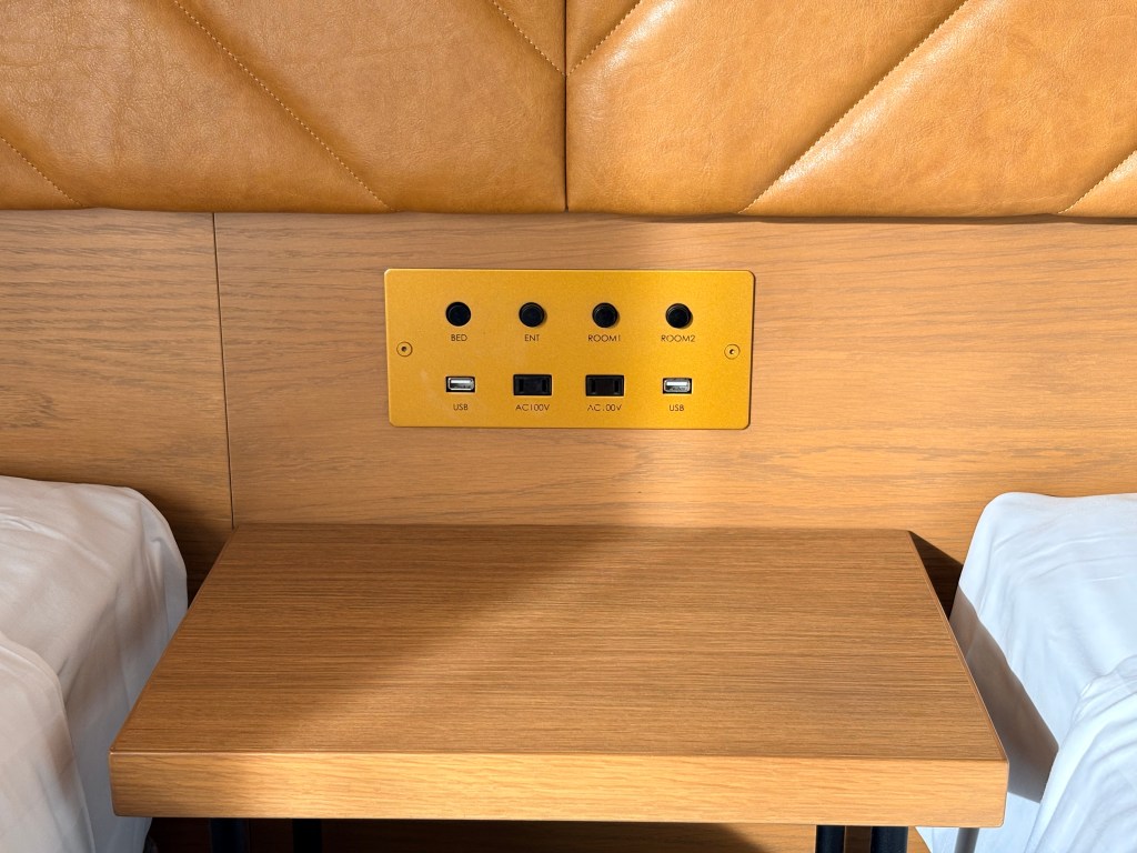 A close-up of the wooden headboard panel between the beds, featuring two USB ports, two AC power outlets, and four light switches.