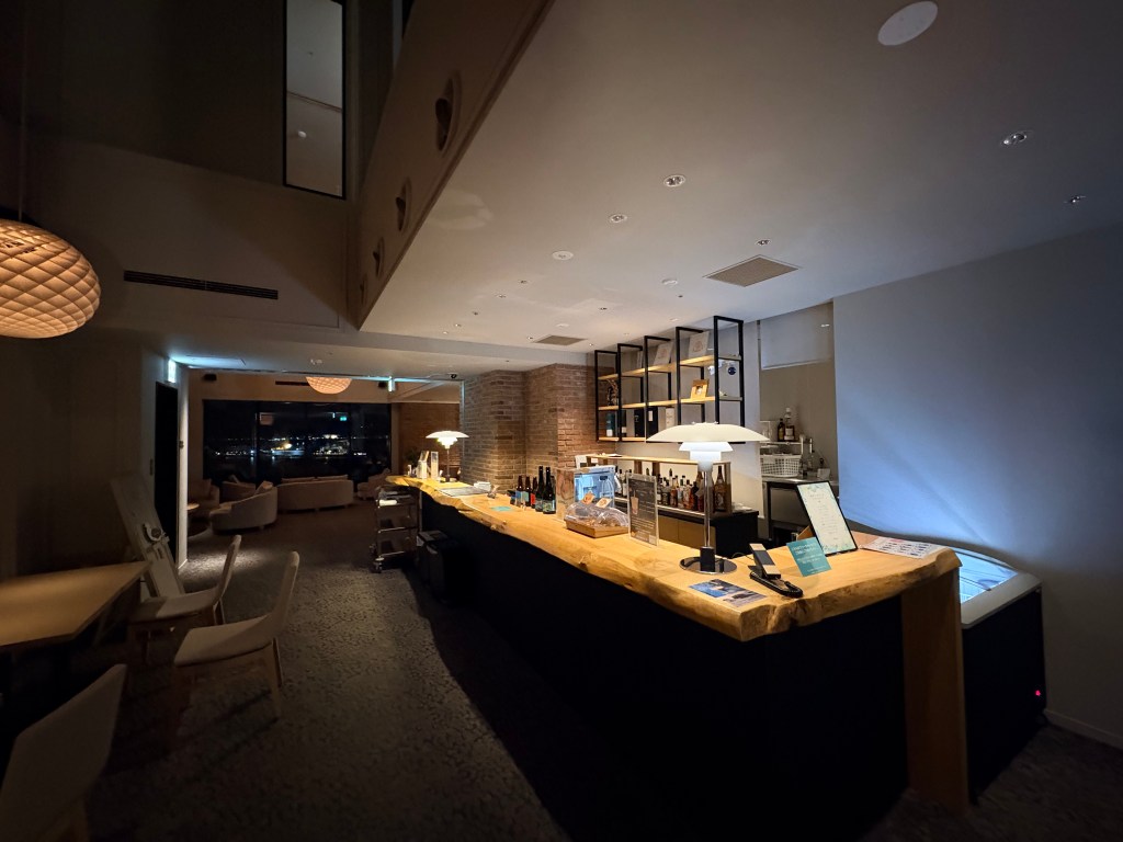 Interior of the modern, warm-lit hotel lounge on the 18th floor at night, featuring dark wood paneling, contemporary seating areas, and large windows looking out at blurred city lights.