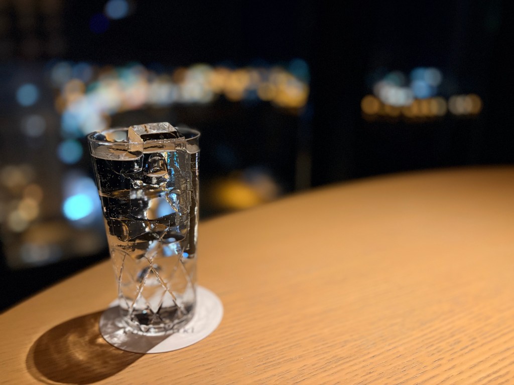 A glass of beer and a glass of shochu sitting on a high wooden table in the lounge, positioned perfectly to frame the dark, majestic silhouette of Mt. Hakodate across the bay through the large windows.