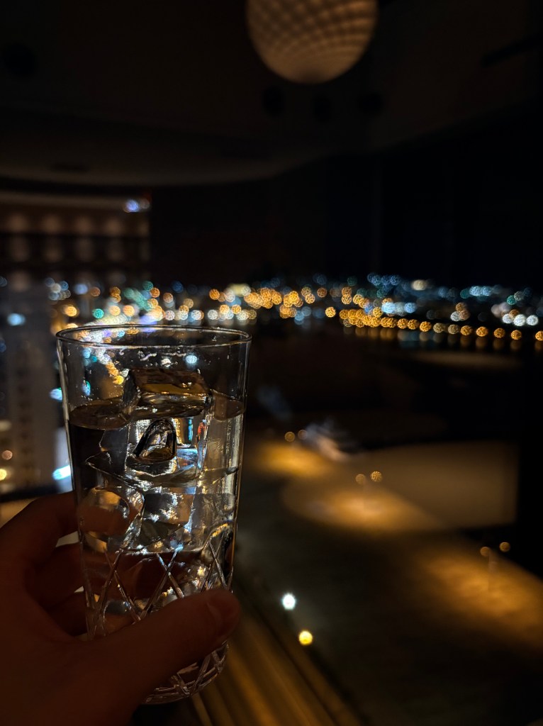 A close-up shot focusing on two drinks on a wooden table, with a slightly blurred but recognizable panoramic view of the sparkling city night lights of Hakodate stretching out below the windows.