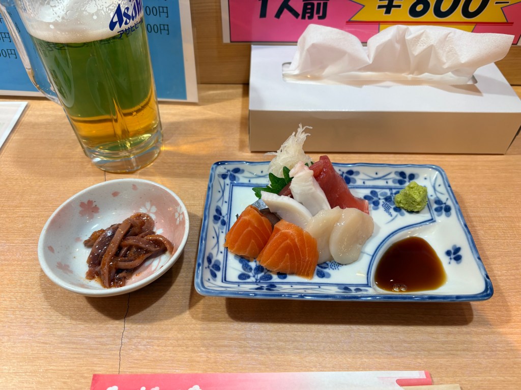 A glass of draft beer next to a small plate of assorted fresh sashimi and a side of salted squid guts on a wooden counter.
