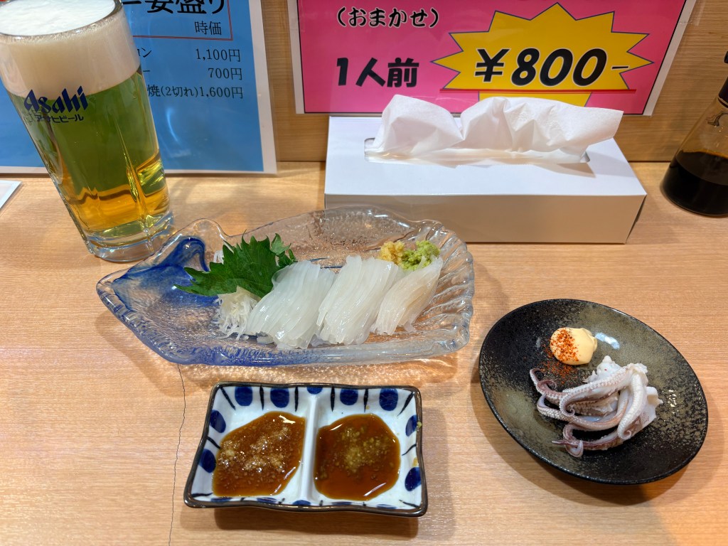 Freshly sliced translucent squid sashimi served on a glass plate with ginger and wasabi, accompanied by grilled squid tentacles and a glass of beer.
