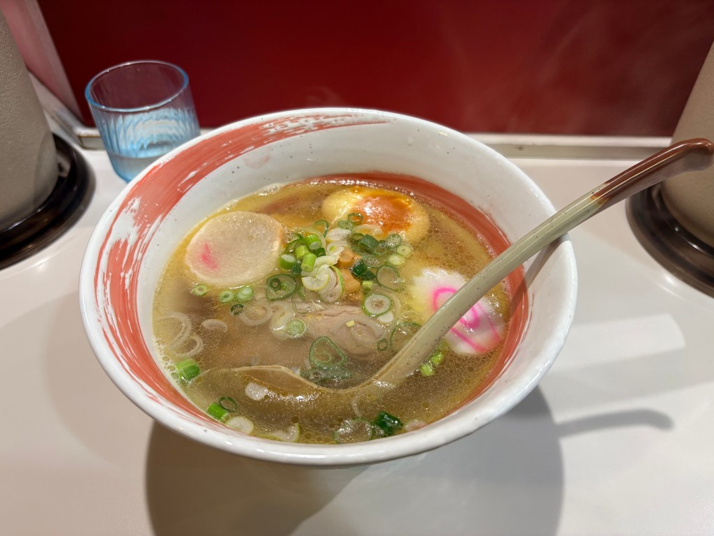 A close-up photograph of a bowl of Hakodate-style Shio (salt) ramen. It features thin, curly yellow noodles in a crystal-clear broth, topped with chashu pork, bamboo shoots, green onions, nori seaweed, naruto fish cake, and a piece of fu (wheat gluten).