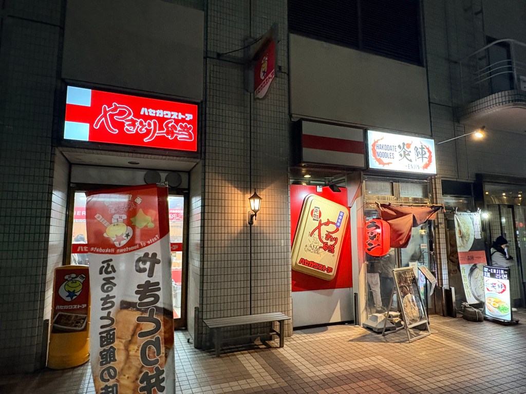 nighttime photograph of the storefront of Hakodate Menya Enjin, featuring a red sign and white paper lanterns. It is located directly adjacent to a brightly lit yellow Hasegawa Store sign.