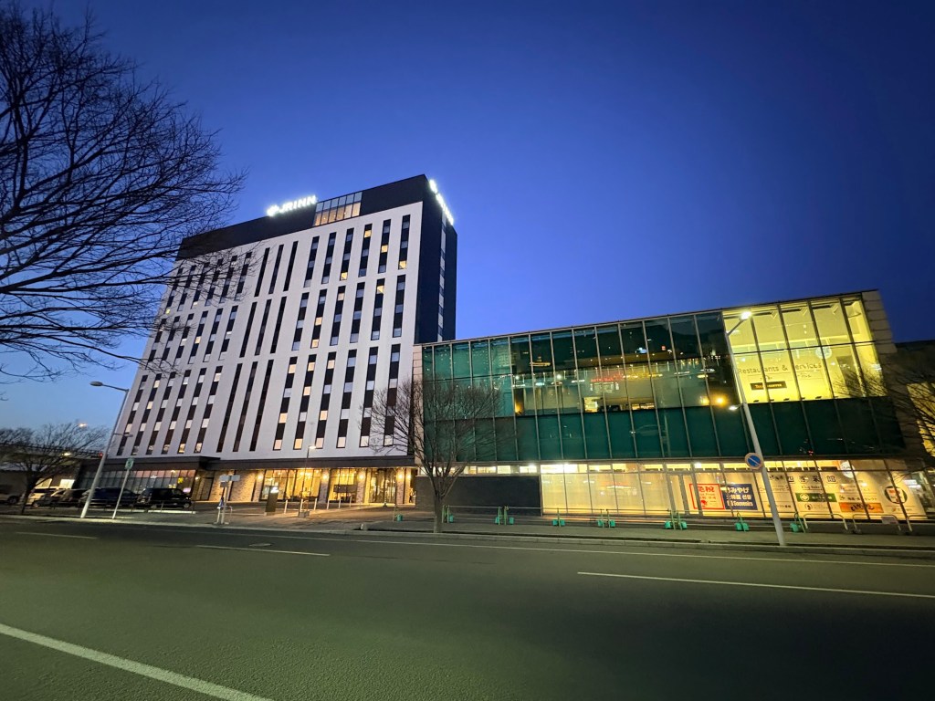 A night view photograph of the modern, lit-up building of JR INN Hakodate, situated directly adjacent to the Hakodate Station building under a dark sky.