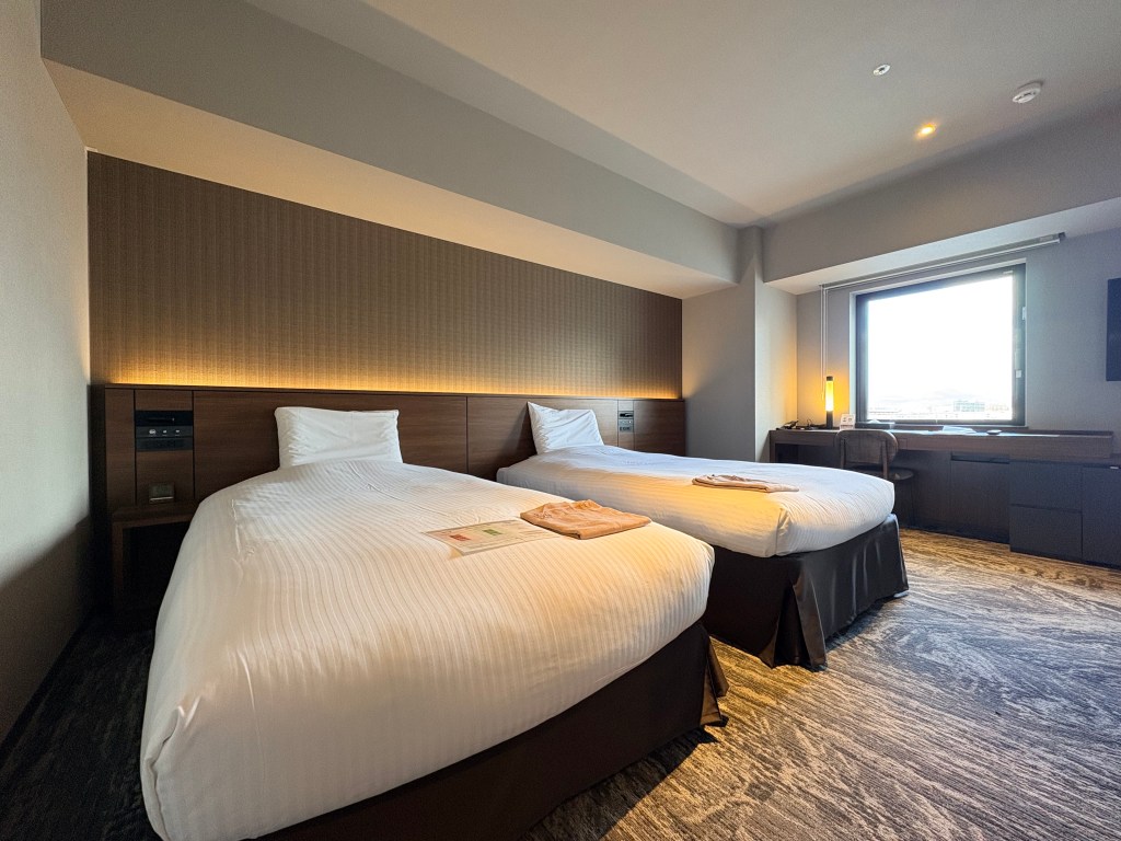 A wide view of a modern hotel premier twin room, showing two beds, a small sofa, table, TV, and a large window looking out at train tracks.