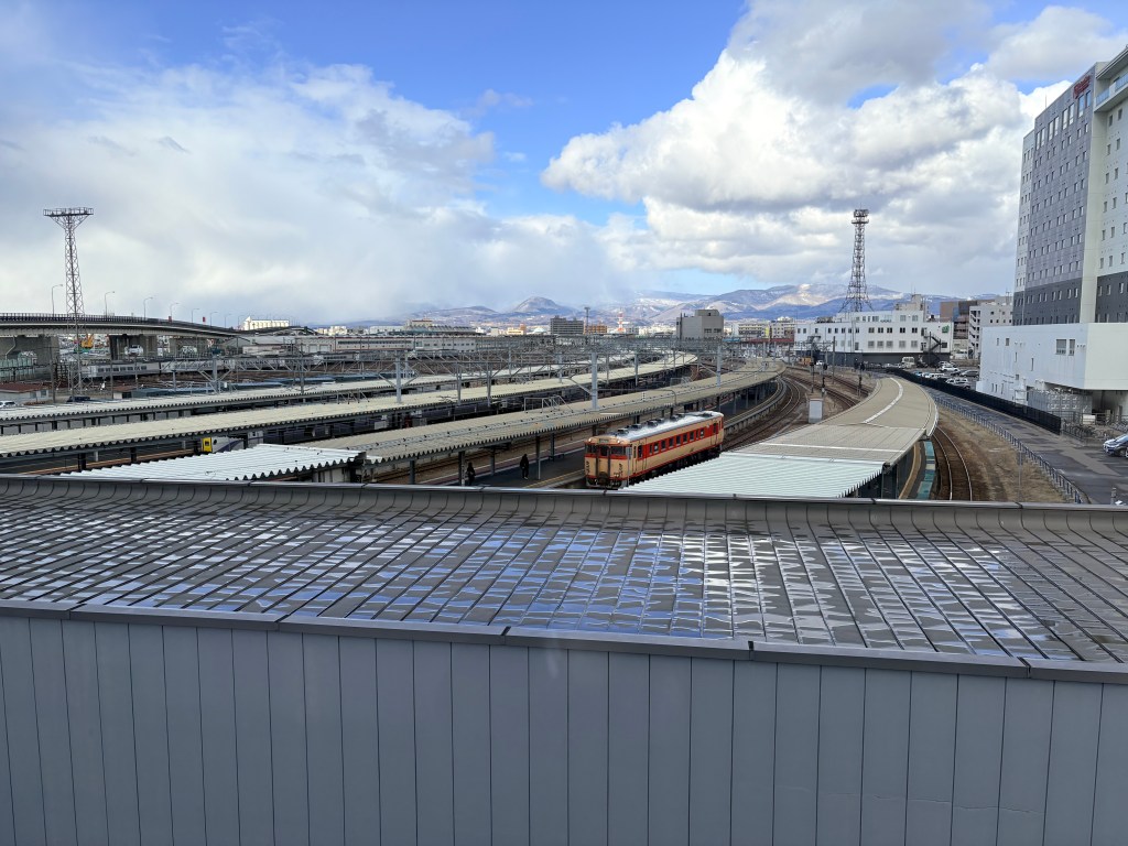 A high-angle view from a guest room window looking down at Hakodate Station's platforms and tracks with a classic red and cream train, set against a backdrop of distant mountains and a blue sky.