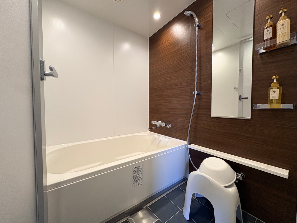 A clean, modern hotel bathroom interior featuring a large, deep white bathtub with a shower head and separated washing area.