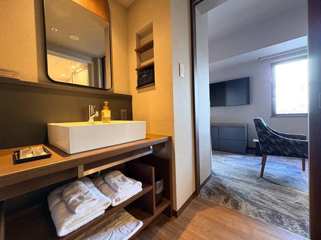 A focused shot of a modern hotel washroom sink area, featuring a white basin, a large mirror, soap dispenser, and two cups.