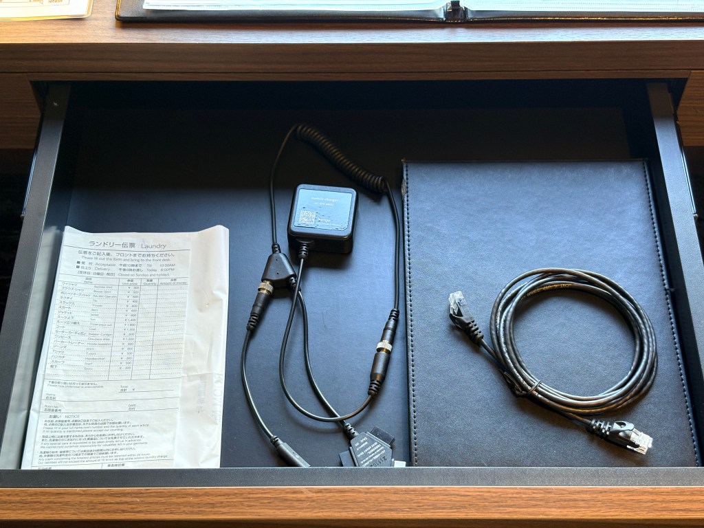 An open desk drawer containing a multi-device charging cable, a black LAN cable, and a laundry service order form.