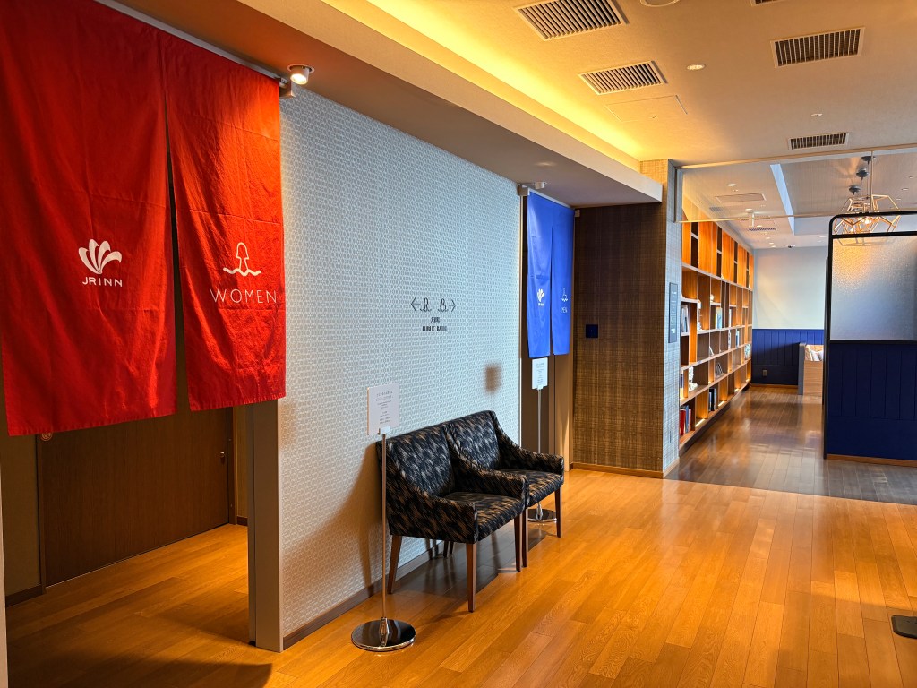 The entrance area of the public hot spring bath showing red curtains for women and blue curtains for men on a clean wooden floor leading into a lounge area.
