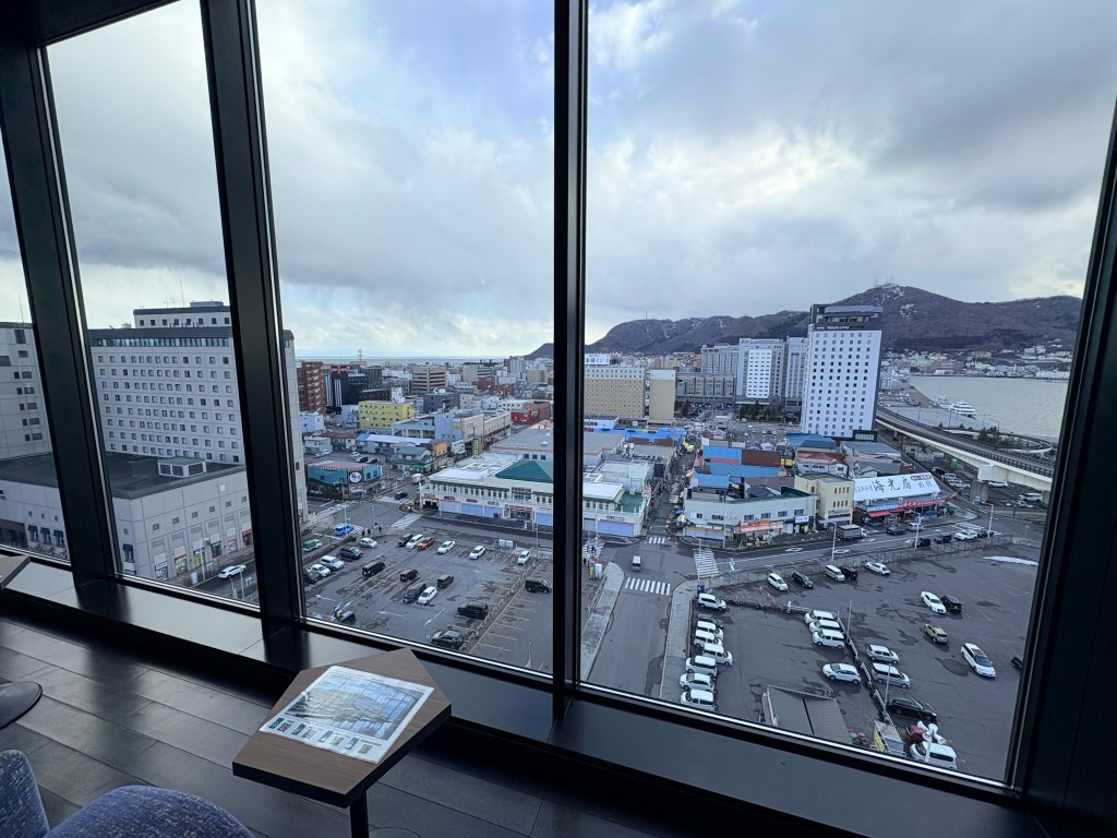 A wide panoramic view through floor-to-ceiling windows in the lounge showing the city of Hakodate, the harbor, and the iconic Mt. Hakodate.