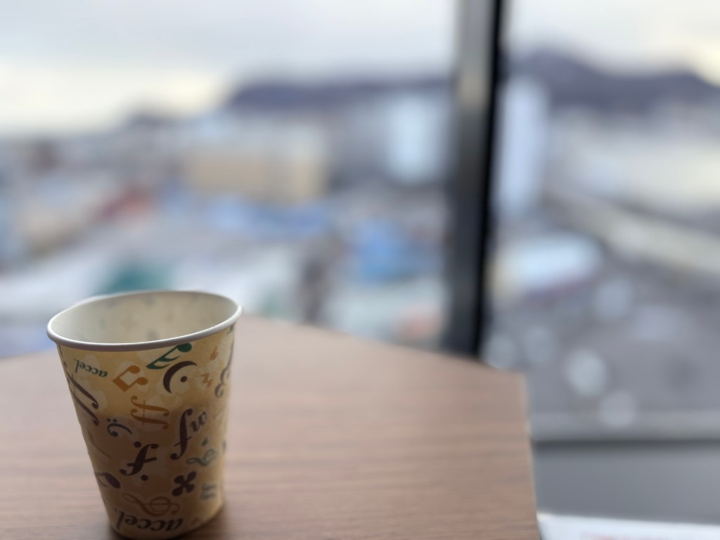 A close-up shot of a decorative paper cup on a wooden table in the lounge, with a blurred view of Mt. Hakodate in the background.