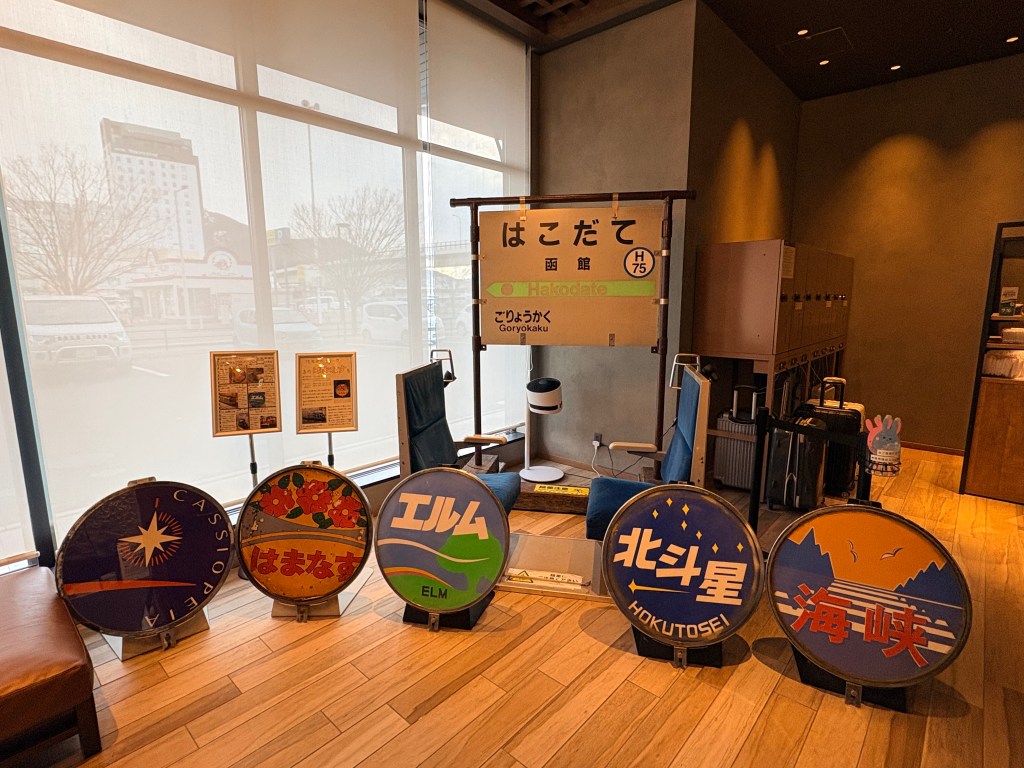 A corner of the hotel lobby featuring various historic colorful train headmarks, a station sign for Hakodate, and vintage train seats.