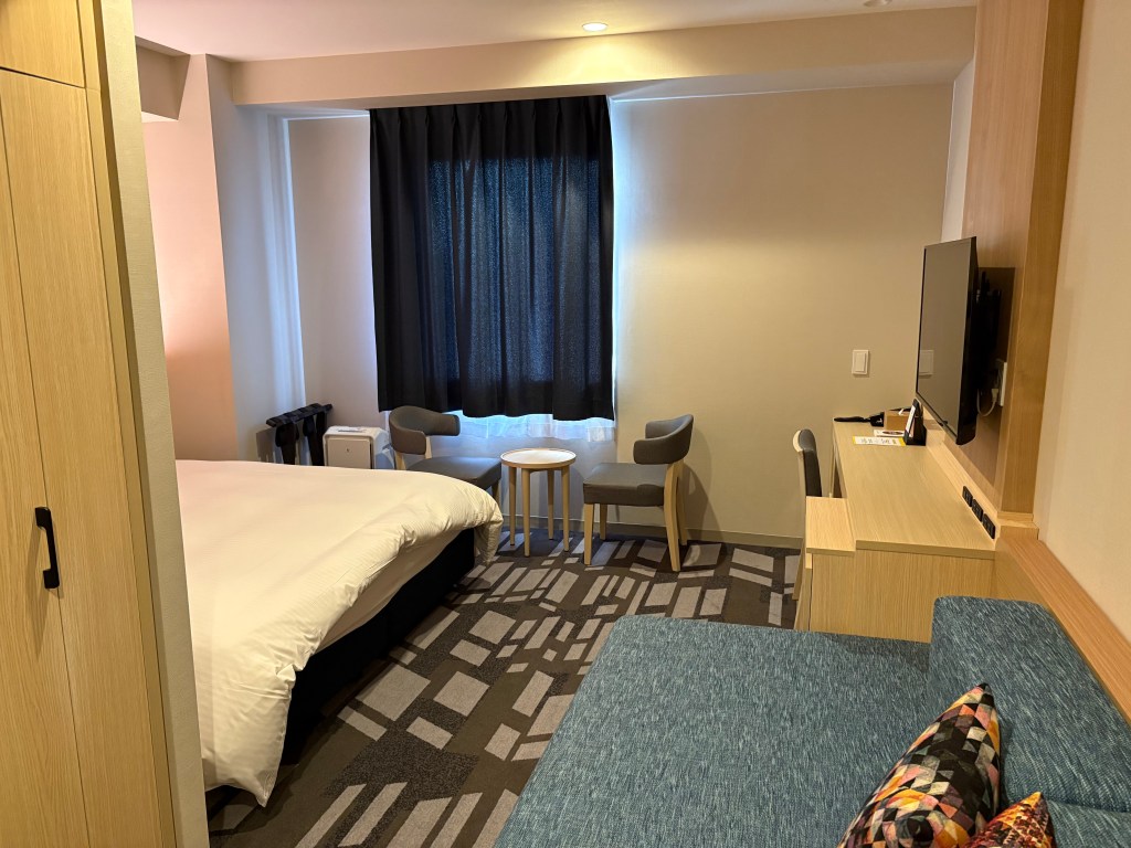 A wide-angle view of a modern hotel premier king room with a large bed, a sofa area, and a functional desk under a window with a city view.