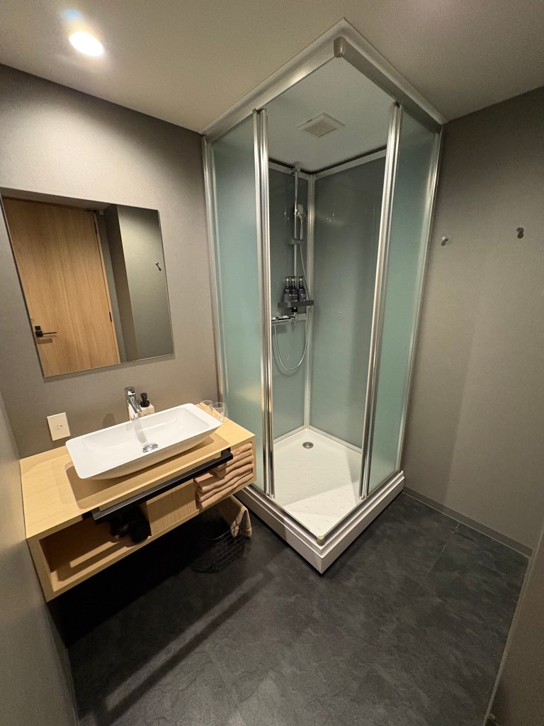 A photograph showing a clean, modern bathroom interior with a single basin sink, a large mirror, and a glass-enclosed shower stall.