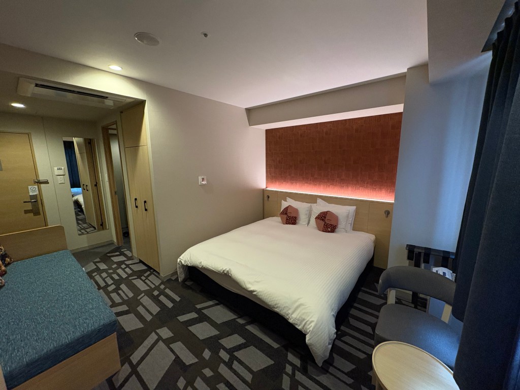 A daytime photograph of a spacious hotel twin room, focusing on two beds with blue runners, a small sofa, and a large window looking out at Hakodate harbor.